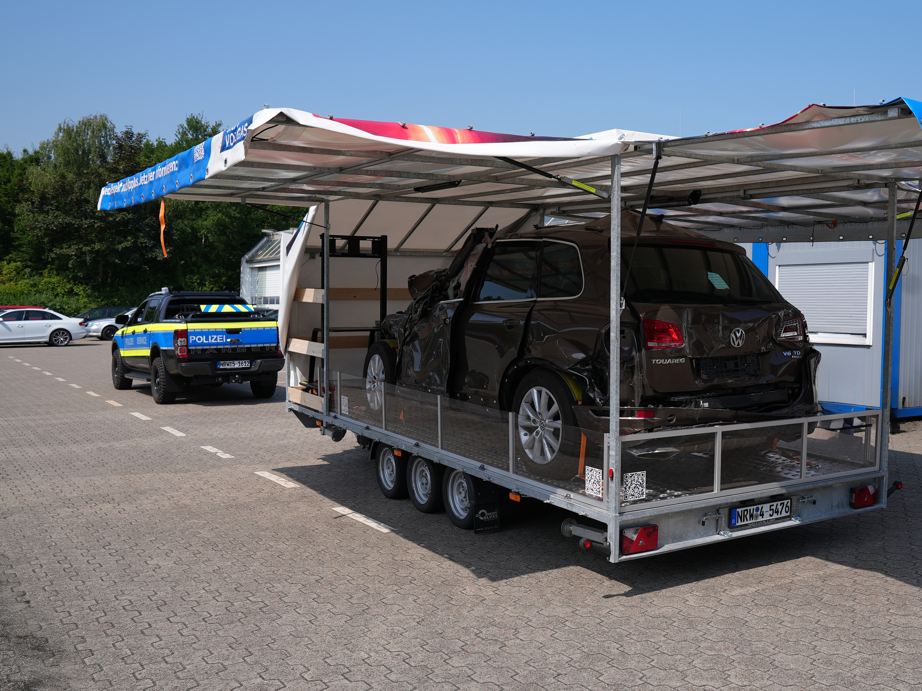 Kampagnenfahrzeug von schräg hinten aufgenommen. Die Seitenwände sind hochgeklappt. Zu sehen ist ein Fahrzeug mit Totalschaden