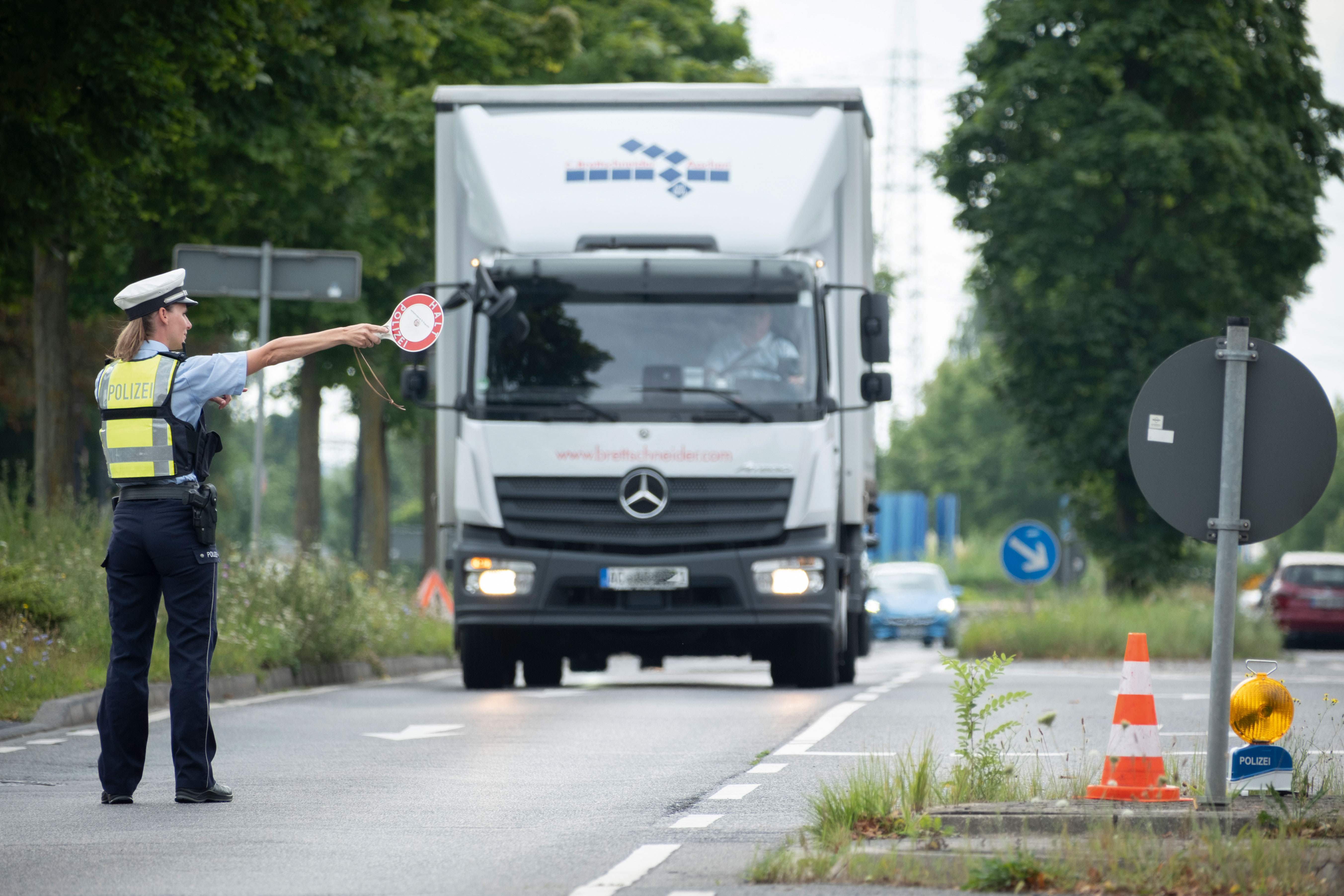 Links, uniformierte Polizistin mit Polizeimütze auf dem Kopf und rot-weißer Anhaltekellein der rechten Hand. Sie steht auf der Straße. Auf sie rollt ein weißer LKA mit blauem Logo oberhalb des Führerhauses zu. Ihr Arm mit Anhaltekelle zeigt nach rechts auf einen Parkplatz.